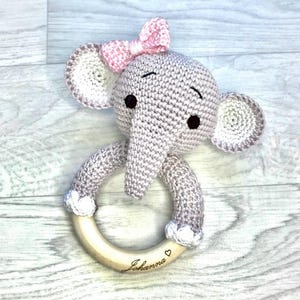 Könnte beinhalten: Ein gehäkeltes Elefanten-Babyrassel mit einem Holzring. Der Elefant ist grau mit schwarzen Augen und einer rosa Schleife. Der Holzring ist mit dem Namen "Johanna" und einem Herz graviert. Ein bezauberndes Babyspielzeug.