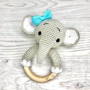 Könnte beinhalten: Handgehäkeltes Elefanten-Babyrassel in Grau mit hellblauer Schleife. Die Rassel hat einen Holzringsockel mit dem Namen "Toni" und einem eingravierten Herz. Der Elefant hat weiße und graue Ohren.