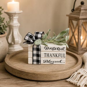 May include: A decorative stack of three white wooden blocks with the words "Grateful, Thankful, Blessed" in black script. A black and white checkered ribbon and greenery adorn the top. The blocks sit on a round wooden tray.