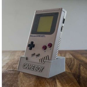 May include: A gray Game Boy handheld video game console is displayed in a gray stand. The console has a black and white design with a gray screen and a red button. The stand has the words "GAME BOY" printed on the front.
