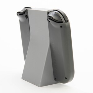 May include: A gray plastic stand designed to hold a gaming console controller. The stand has a triangular base and a vertical section to support the controller. The controller is dark gray with black buttons.