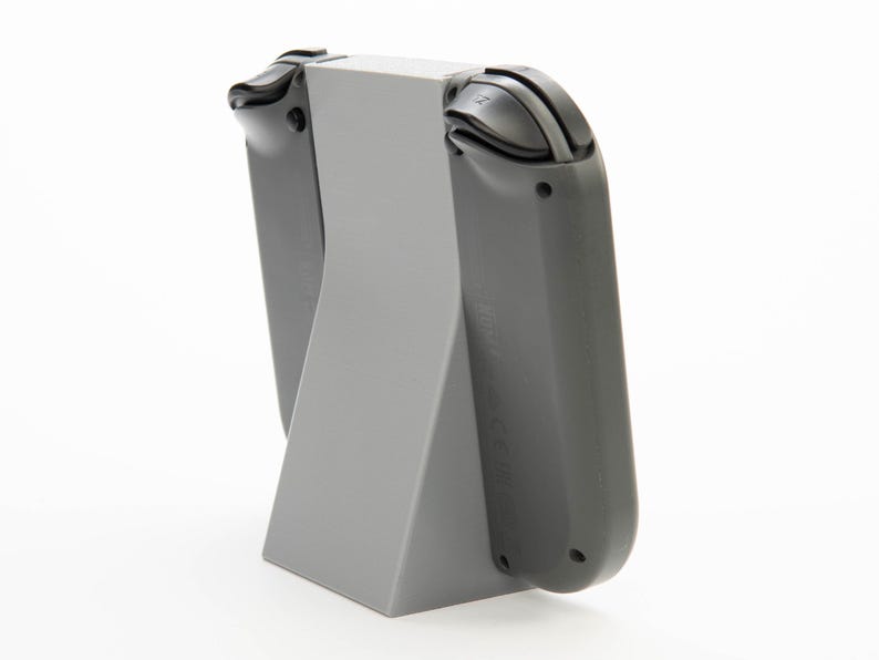 May include: A gray plastic stand designed to hold a gaming console controller. The stand has a triangular base and a vertical section to support the controller. The controller is dark gray with black buttons.