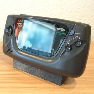 May include: A black Sega Game Gear handheld video game console with a grey screen and a stand. The console has a speaker, a start button, and a volume control knob. The text "SEGA PORTABLE VIDEO GAME SYSTEM" is printed on the console.