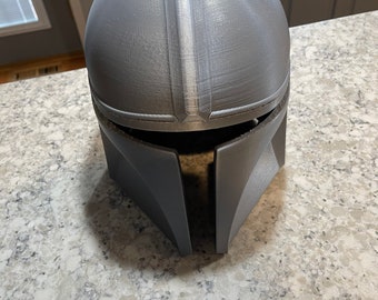 May include: A grey Mandalorian helmet, possibly 3D-printed, with a smooth, matte finish. The helmet has a distinctive T-shaped visor and angular cheek plates. A vertical ridge runs along the top. A cosplay or collectible item.