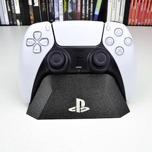 May include: A white PlayStation 5 DualSense controller is sitting on a black stand with the PlayStation logo. The controller has black buttons and analog sticks.