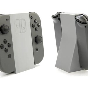 May include: Two gray Nintendo Switch Joy-Con controllers with a light gray center section, one attached to a light gray stand. The controllers have black buttons and joysticks. The stand is angled for display.