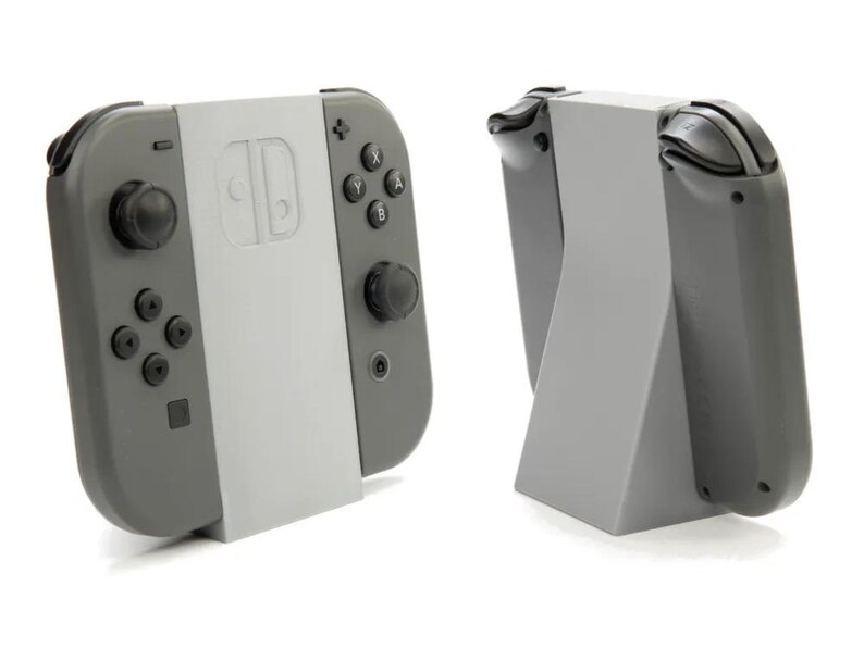 May include: Two gray Nintendo Switch Joy-Con controllers with a light gray center section, one attached to a light gray stand. The controllers have black buttons and joysticks. The stand is angled for display.