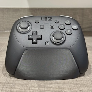 Puede incluir: Un mando Nintendo Switch Pro negro con detalles blancos. El mando tiene una cruceta, palancas analógicas y botones de acción. El logotipo de Nintendo Switch es visible. Diseñado para jugar cómodamente.