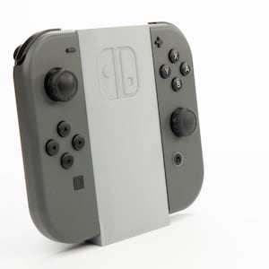 May include: A gray Nintendo Switch Joy-Con controller with a light gray stand. The controller has black buttons and joysticks. The stand has the Nintendo Switch logo.