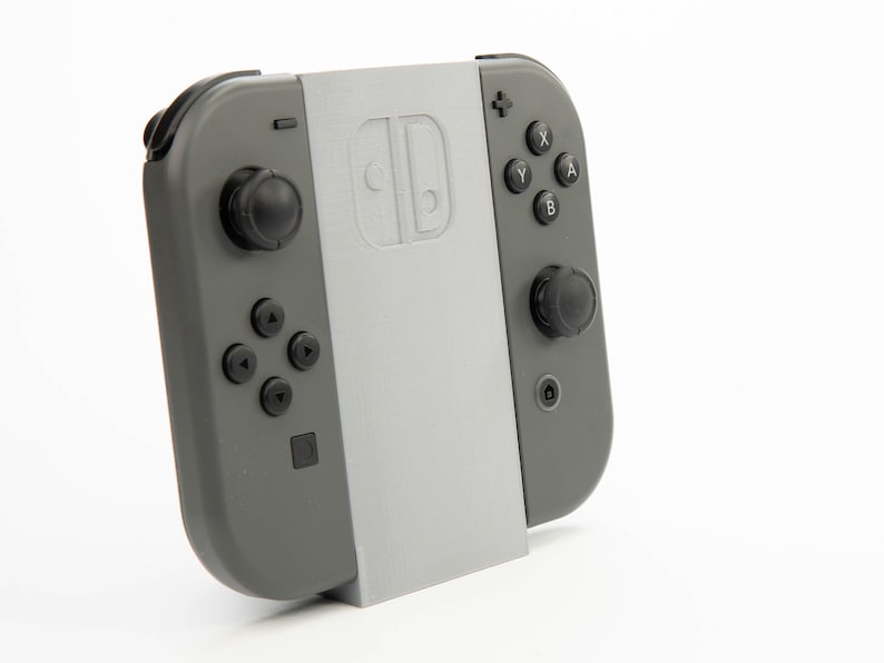 May include: A gray Nintendo Switch Joy-Con controller with a light gray stand. The controller has black buttons and joysticks. The stand has the Nintendo Switch logo.