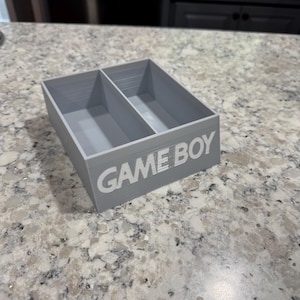 May include: A gray plastic storage box with two compartments. The box has the words "GAME BOY" printed on the front in white.