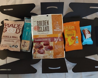 May include: A black box with five different snack bags inside. The bags are labeled "Made Good Granola Minis Chocolate Chip", "Golden Island Snack Bites Korean Barbecue", "Solely Organic Mango Orange Whole Fruit Gummies", and "Unreal Dark Chocolate Peanut Butter Cups".
