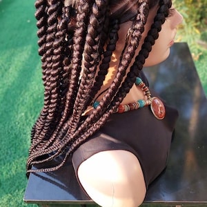 May include: A brown wig with long, thick braids. The wig is styled in a way that the braids are pulled back and away from the face. The wig is on a mannequin head and is sitting on a black table.