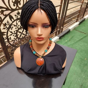 May include: A black braided wig with a lace front, worn on a mannequin head. The mannequin is wearing a black top and a multi-coloured beaded necklace with a large, round, red pendant.