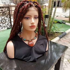 Puede incluir: Una peluca marrón con extensiones largas trenzadas. La peluca está puesta en una cabeza de maniquí con una blusa negra y un collar de cuentas marrones y turquesas.