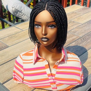 Könnte beinhalten: Eine schwarze geflochtene Perücke mit einem Spitzenfront. Die Perücke ist in einem kurzen Bob mit kleinen Zöpfen gestylt. Die Perücke befindet sich auf einem Mannequin-Kopf, der ein gestreiftes Hemd trägt.