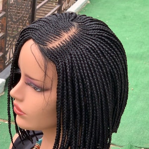 Puede incluir: Una peluca negra trenzada con un corte bob, exhibida en una cabeza de maniquí. La peluca presenta pequeñas trenzas cuidadosamente tejidas y una línea de cabello de aspecto natural. El maniquí lleva un collar con cuentas marrones, naranjas y turquesas.