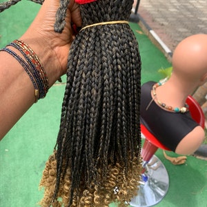 May include: A bundle of black and brown braided hair extensions with curly, light brown ends. The braids are secured with a rubber band and red thread. The hair is held in a hand, with beaded bracelets visible on the wrist.