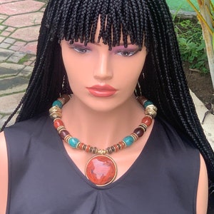 May include: A mannequin head with a black braided wig and a statement necklace. The necklace features large, round beads in shades of red, brown, turquoise, and gold. A large, round pendant with a reddish-brown stone hangs from the necklace.