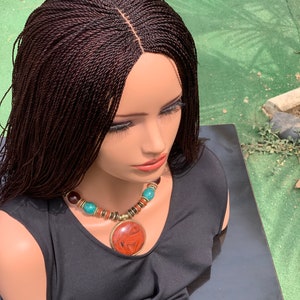 May include: A brown wig with small braids, styled in a long, straight style. The wig has a lace front and is worn on a mannequin head. The mannequin is wearing a black tank top and a necklace with turquoise and brown beads.