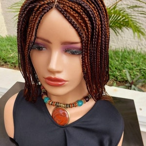 Puede incluir: Cabeza de maniquí que muestra una peluca trenzada con tonos marrones y rojizos. Las trenzas están bien hechas, y la peluca tiene una raya natural. El maniquí lleva una camiseta negra y un collar con un colgante grande y redondo.