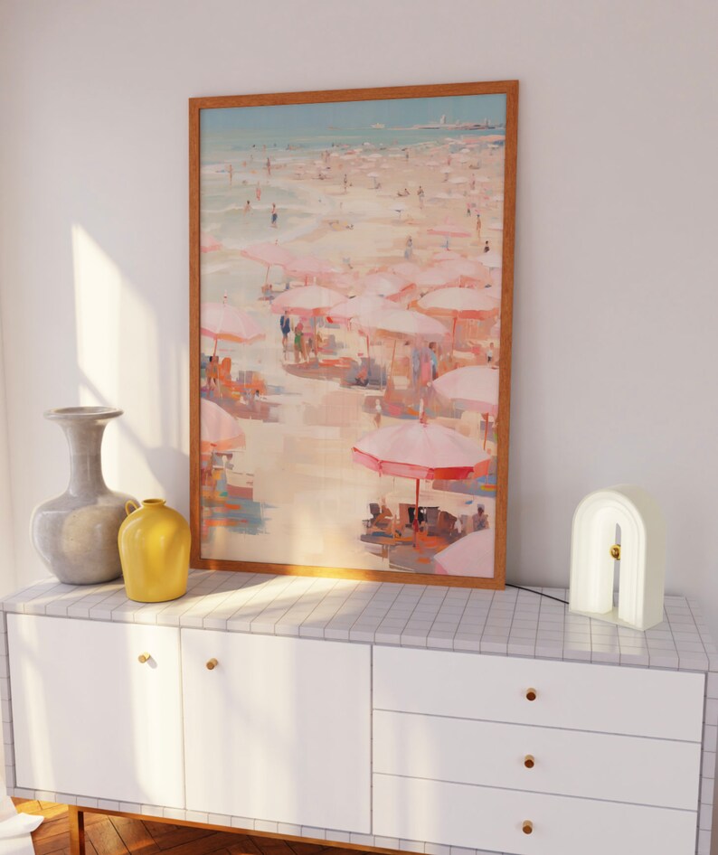 May include: A framed painting of a beach scene with pink umbrellas and people on the sand. The painting is hanging on a white wall above a white dresser with gold knobs.