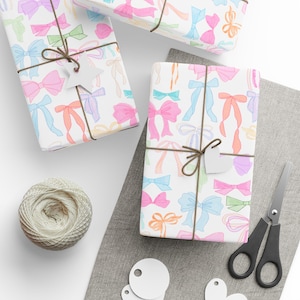 May include: Three gift boxes wrapped in white paper with a colorful bow pattern. The boxes are tied with twine and have blank gift tags attached. The boxes are on a white surface with a gray fabric surface in the background.