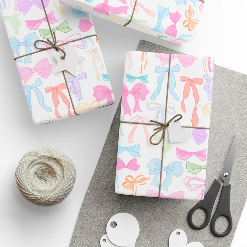 May include: Three gift boxes wrapped in white paper with a colorful bow pattern. The boxes are tied with twine and have blank gift tags attached. The boxes are on a white surface with a gray fabric surface in the background.