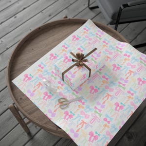 May include: A white gift box wrapped in colorful bow patterned wrapping paper. The box is tied with a brown ribbon and sits on a wooden tray.
