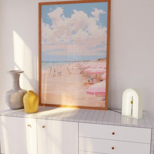 May include: A white dresser with three drawers and gold knobs. A framed print of a beach scene with pink umbrellas hangs above the dresser. A white arch-shaped lamp with a gold accent sits on the dresser.