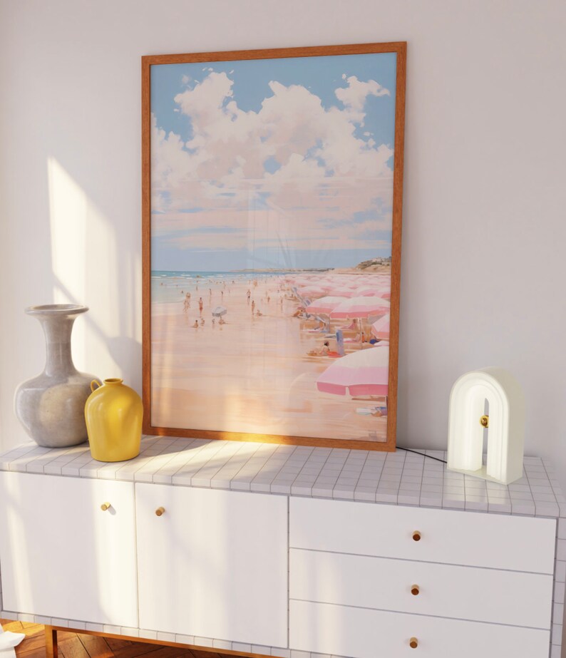 May include: A white dresser with three drawers and gold knobs. A framed print of a beach scene with pink umbrellas hangs above the dresser. A white arch-shaped lamp with a gold accent sits on the dresser.