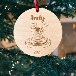 May include: A round wooden ornament with a red ribbon for hanging. The ornament features the name "Andy" above an illustration of hands shaping a vase on a pottery wheel. The year "2025" is etched below the illustration.