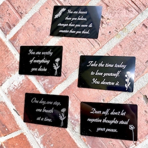 May include: Five black metal cards with inspirational quotes in white script. The cards feature floral accents and phrases like "You are braver than you believe" and "You are worthy of everything you desire."