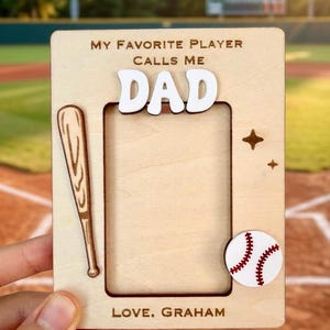 May include: A rectangular light wood picture frame featuring the engraved text MY FAVORITE PLAYER CALLS ME DAD and LOVE, GRAHAM. The frame includes a raised white DAD cutout, a wooden baseball bat graphic, a white baseball, and two small stars.