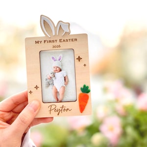 May include: Wooden photo frame with bunny ears, the text "MY FIRST EASTER 2026" and a carrot. The frame holds a baby photo. The baby is wearing a bunny hat and holding a stuffed animal. The name "Peyton" is written on the frame.