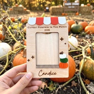 May include: A wooden photo frame with a red and white awning design at the top. The frame has the text "Cutest Pumpkin in the Patch 2026" and the name "Candice". The frame is decorated with star shapes and stacked pumpkin cutouts.
