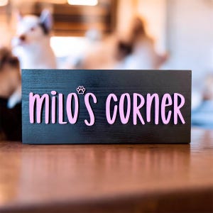 May include: A rectangular black wooden sign featuring the light pink text "milo's corner" in a playful font. A small light pink paw print icon is positioned above the letter i in the word milo's, creating a decorative accent on the surface.