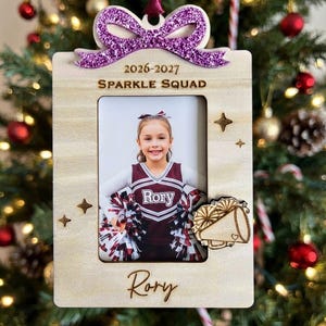 May include: A rectangular wooden photo frame ornament featuring a purple glitter bow at the top, engraved with the text 2026-2027, SPARKLE SQUAD, and the name Rory. The frame includes a photo of a cheerleader and a small wooden megaphone cutout.