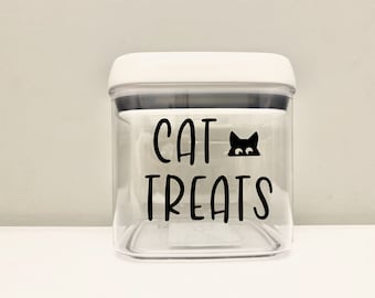 May include: A clear, square container with a white lid, labeled "CAT TREATS" in black script. A black cat silhouette peeks from behind the text. The container is designed for storing cat treats.