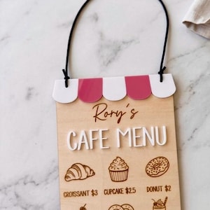 May include: A rectangular wooden cafe menu sign with a black hanging cord and a pink and white scalloped awning top. The sign features the text Rory's CAFE MENU and engraved illustrations of a croissant, cupcake, donut, cake slice, pretzel, and milkshake.