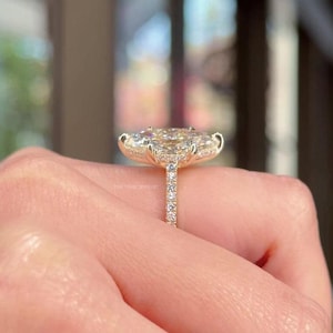 May include: A gold ring with a large, oval-cut diamond surrounded by a halo of smaller diamonds. The band is also set with small diamonds. The ring is held up by a hand. The text "THE TRILL JEWELRY" is visible.