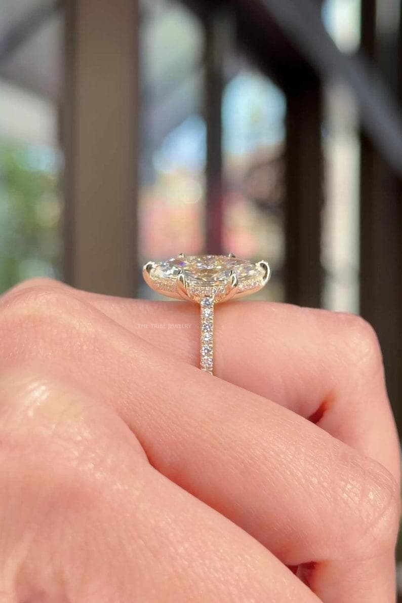May include: A gold ring with a large, oval-cut diamond surrounded by a halo of smaller diamonds. The band is also set with small diamonds. The ring is held up by a hand. The text "THE TRILL JEWELRY" is visible.