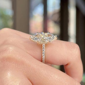 May include: A gold engagement ring with a large, oval-cut diamond and a halo of smaller diamonds. The band is set with small diamonds. The ring is held by a hand.