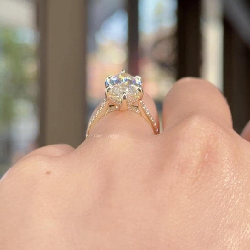 May include: A gold engagement ring with a large, clear, oval-cut gemstone. The ring has a delicate band with small accent stones. The words "THE TRIBE JEWELS" are visible on the ring's band.