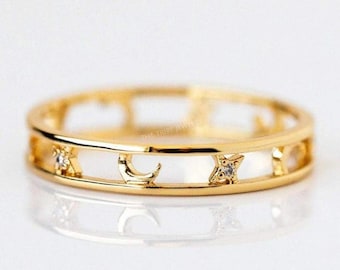 May include: A gold-colored ring with celestial designs, including a crescent moon, a star, and a four-pointed star, each with a small, clear gemstone. The ring is on a white surface and has the text "THE TRIBE JEWELS".