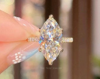 May include: A marquise-cut diamond engagement ring with a gold band. The band is encrusted with small diamonds. The ring is held up in front of a blurred background. The text "THE TRIBE JEWELRY" is visible.