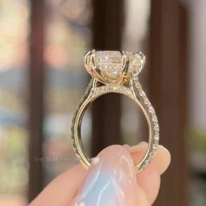 May include: A gold engagement ring featuring a large, clear, oval-cut central diamond and smaller accent stones. The band is also embellished with small, sparkling stones. The ring is held up, displaying its intricate design. The Tribe Jewelry is printed on the ring.