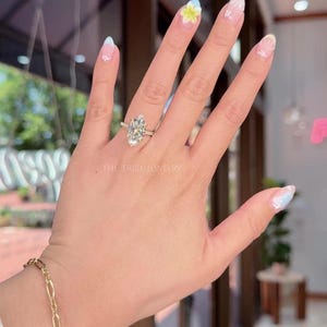 May include: A hand displaying a silver ring with a large, pear-shaped diamond. The ring is set on a delicate band. The text "THE TRIBE JEWELRY" is visible. A gold bracelet is also visible.