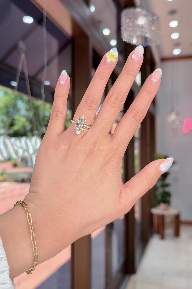 May include: A hand displaying a silver ring with a large, pear-shaped diamond. The ring is set on a delicate band. The text "THE TRIBE JEWELRY" is visible. A gold bracelet is also visible.