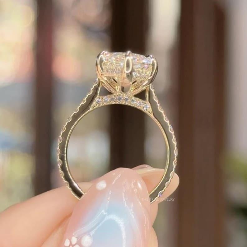 May include: A gold engagement ring featuring a large, clear, cushion-cut diamond. The band is embellished with smaller diamonds. The ring is held up by a hand, highlighting the intricate design and the sparkle of the gemstones.